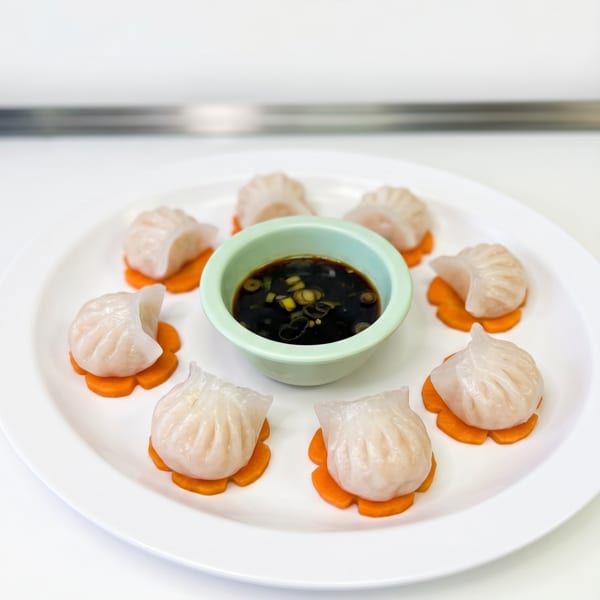 Seven steamed dumplings with translucent wrappers on carrot slices, surrounding a bowl of soy sauce.