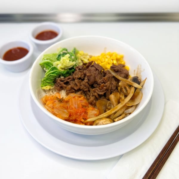 Bibimbap bowl with beef, kimchi, corn, and greens, served with two sauce dishes and chopsticks.
