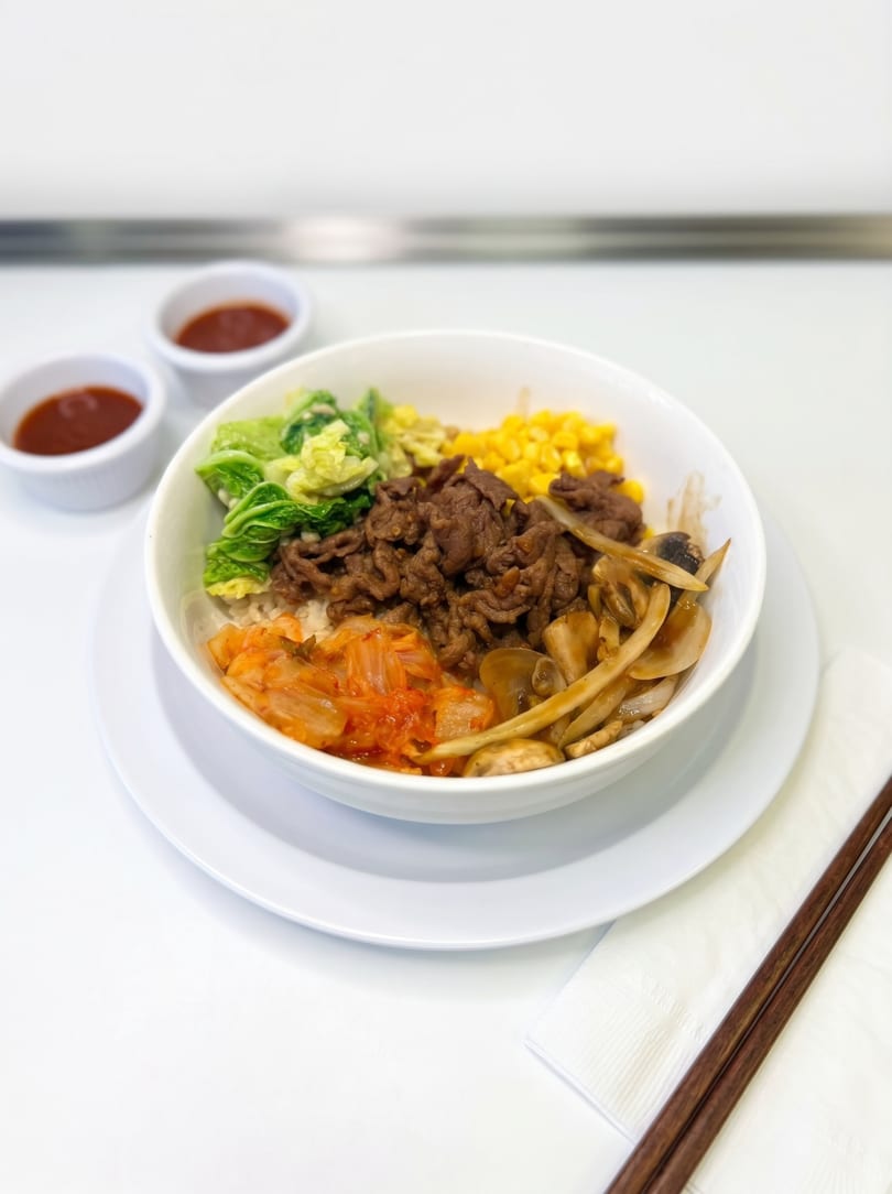 Bibimbap bowl with beef, kimchi, corn, and greens, served with two sauce dishes and chopsticks.